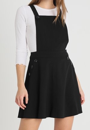 Sort overall-kjole med justerbare stropper, der har store knapper på siderne og et skørt med udstående design lavet af glat stof.