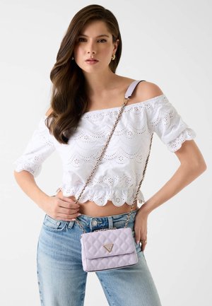 Femme portant un chemisier blanc à broderies anglaises décolleté, un jean bleu clair et un petit sac bandoulière matelassé lavande avec une chaîne dorée.