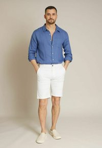 Chemise en lin bleue avec des fermetures à boutons, shorts en coton blancs et chaussures slip-on beiges. Le modèle se tient avec les mains dans les poches sur un fond neutre.