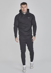 Black hoodie and pants set made of smooth fabric. Hoodie features a front pocket, ribbed cuffs, and a small logo on the chest. Silver sneakers.