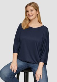 Glimlachende vrouw met lang blond haar, gekleed in een navyblauwe losse top en blauwe jeans, zittend op een zwarte kruk tegen een lichte achtergrond.
