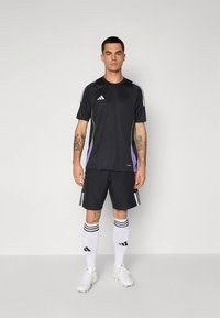 Must mustriga lühikeste varrukatega sportlik särk, millel on valged ja lillad aktsendid, sobitatud mustade lühikeste pükstega ning valgete triibuliste sokkidega.