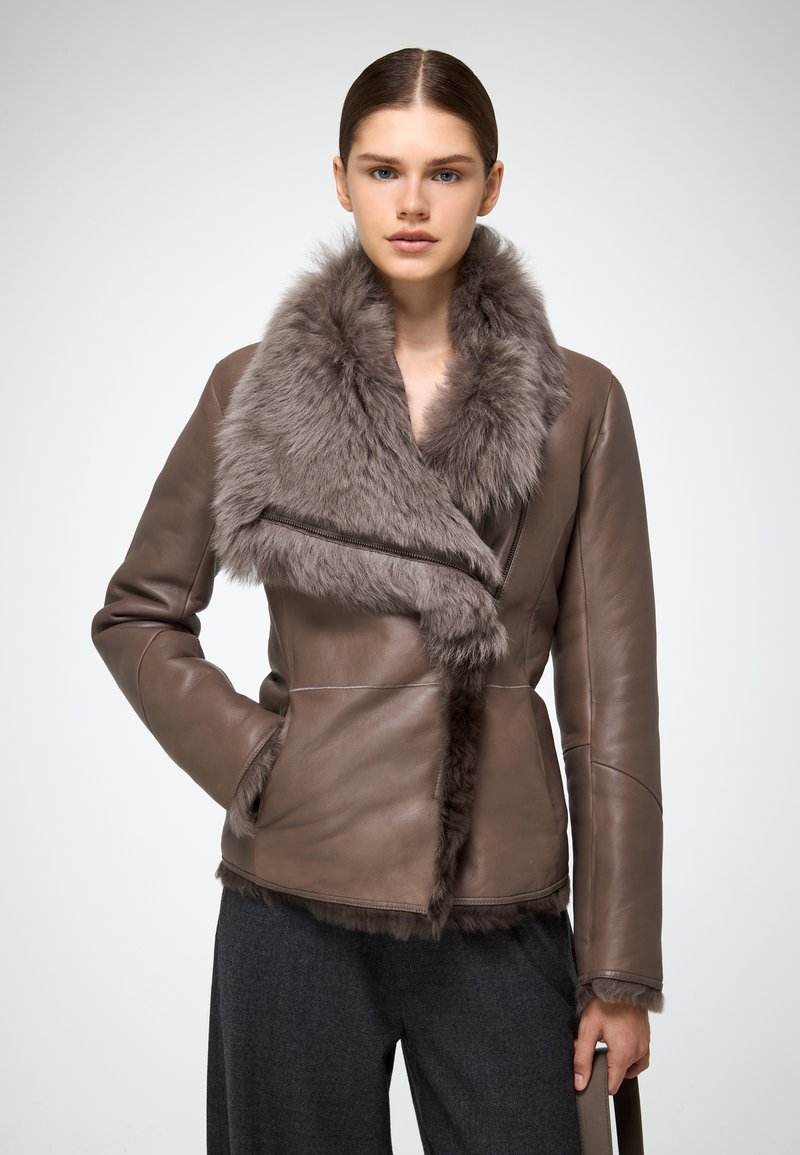Giacca di pelle marrone con collo in pelliccia ecologica, design asimmetrico e silhouette slanciata. Presenta chiusura con zip e texture morbida.