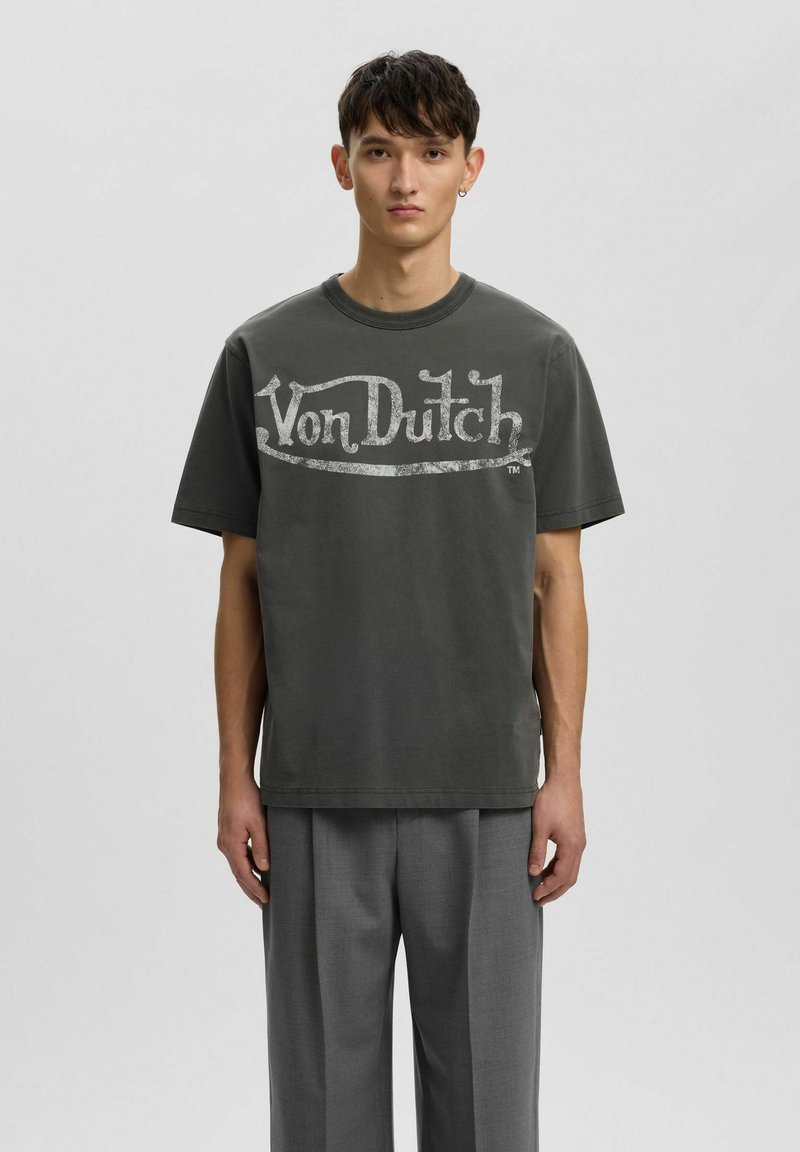 Jeune homme aux cheveux courts et foncés, portant un t-shirt gris foncé avec le logo "Von Dutch" et un pantalon gris, debout devant un fond uni.