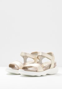 Zlaté metalické sandály se dvěma předními popruhy, zadním popruhem a polstrovanou bílou podrážkou. Mají hladkou texturu a otevřenou špičku.