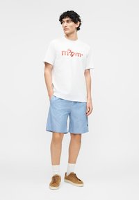 Jeune homme aux cheveux bouclés portant un t-shirt blanc à motif, un short rayé bleu et blanc, des chaussettes blanches et des baskets marron, debout les mains dans les poches.