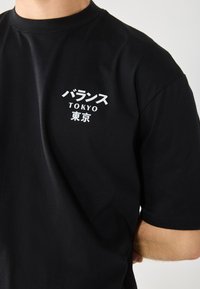 Crna pamučna majica s bijelim tekstom na japanskom i engleskom: "バランス TOKYO." Kratki rukavi i okrugli izrez s opuštenim krojem.