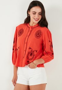 Rote Langarmbluse mit schwarzem floralem Stickereimuster, mit Knopfleiste vorne und gerafften Ärmeln, kombiniert mit weißen Shorts. Leichtes Material.