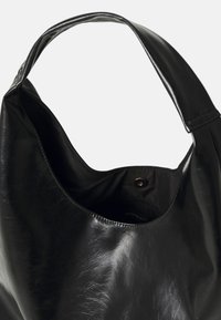 Sac hobo en cuir noir avec une texture lisse, une large bandoulière et un grand dessus ouvert. L'intérieur possède une doublure noire et une poche zippée supplémentaire.