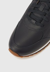 Tamnoplava kožna tenisica s teksturiranim detaljima, zaobljenim vrhom, ravnim vezicama i gumenim potplatom u boji gume. Ima bijele detalje na srednjem potplatu.