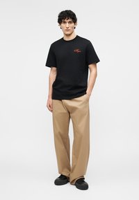 Homme portant un T-shirt noir à manches courtes avec un logo orange, un pantalon beige à jambes larges et des baskets noires, debout devant un fond blanc uni.