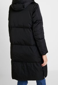 Abrigo negro acolchado con capucha, con un patrón acolchado, mangas largas y una cremallera lateral. Hecho de una tela lisa e aislante.