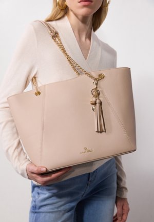 Femme portant un pull crème et un jean tient un grand sac à main en cuir beige avec une bandoulière chaîne dorée et un pompon.