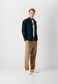 Dunkelgrünes kariertes Hemd über einem weißen T-Shirt, beige Hose und weiße Sneaker mit beigen Akzenten. Glatter Stoff, lässige Passform.