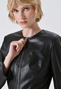 Giacca in pelle nera con colletto rotondo, cuciture visibili e chiusura con zip dorata. Il materiale è liscio e aderente.