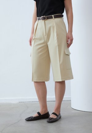 Bermudas beiges avec plis et poches latérales, assorties d'une ceinture marron. Portées avec un haut noir et des ballerines noires.