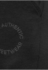 Schwarzer Stoff mit gerippter Textur zeigt ein kreisförmiges, geprägtes Textdesign mit der Aufschrift "AUTHENTIC STREETWEAR."