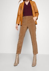Vrouw in tan corduroy broek, beige enkellaarsjes, bordeaux knoopshirt en tan leren blazer, staand met één hand in de zak.
