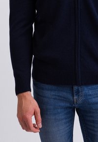 Mörkblå stickad cardigan med dragkedja, ribbade ärmslut, två sidfickor och en strukturerad yta, bärs över blåa denimjeans.