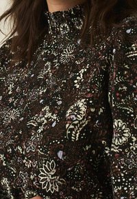 Schwarzbluse mit Blumenmuster und gerafftem Ausschnitt, verziert mit grünen, weißen und lila Blumen auf einem strukturierten Stoff.