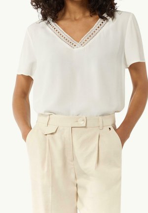 Femme portant un chemisier blanc à manches courtes en V avec bordure en dentelle, associé à un pantalon crème taille haute avec ceinture et poches, mains dans les poches.