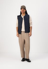 Marineblauwe gewatteerde bodywarmer met geometrische patronen, beige gebreide pullover, losse beige broeken en zwarte schoenen. Grijs en bruin geruit petje inbegrepen.