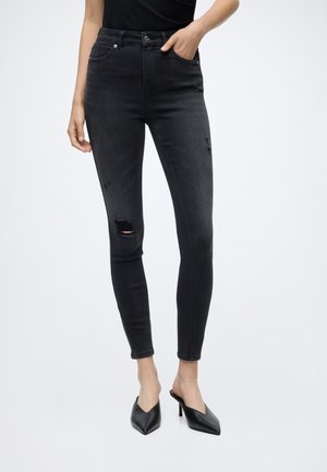Femme portant un jean skinny noir avec des détails usés sur la cuisse et le genou, associé à des mules à talons noires.