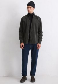 Chemise boutonnée vert foncé sur un col roulé noir, associée à un jean bleu foncé et des chaussures marron. Le modèle porte également un bonnet noir.