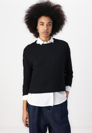 Black cropped sweater with long sleeves, rounded neckline, textured knit, and button accents on shoulders, worn over a white collared shirt.
