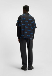 Zwart t-shirt met korte mouwen met een herhalend blauw logo-patroon, gecombineerd met zwarte cargobroek, gezien vanaf de achterkant.