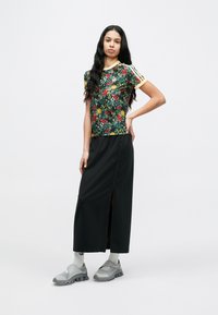 Νεαρή γυναίκα με μακριά μαύρα μαλλιά φοράει ένα φλοράλ κοντομάνικο πουκάμισο, μαύρη μακριά φούστα με σκίσιμο, λευκές κάλτσες και γκρι αθλητικά παπούτσια.