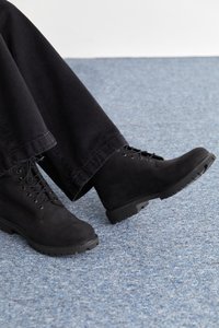 Botas de ante negras con cordones, con una suela resistente y acentos texturizados. Combinadas con pantalones negros holgados sobre una alfombra azul.