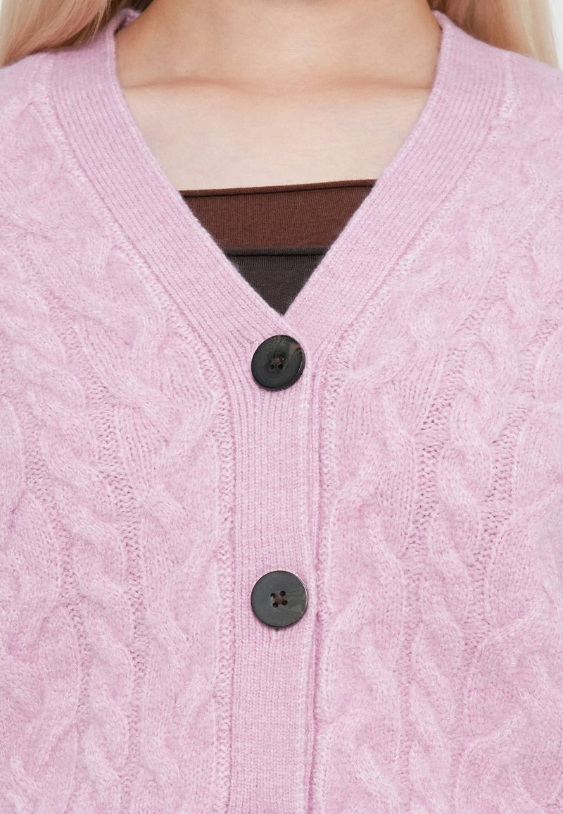 Cardigan en tricot torsadé rose clair avec un col en V, deux boutons noirs et un bord côtelé au niveau de l'encolure et de la patte de boutonnage.