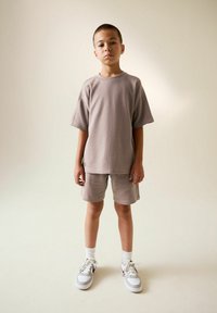 Svetlohnedé bavlnené tričko s krátkymi rukávmi a zodpovedajúcimi šortkami, doplnené o biele tenisky a ponožky. Hladká textúra, voľný strih.