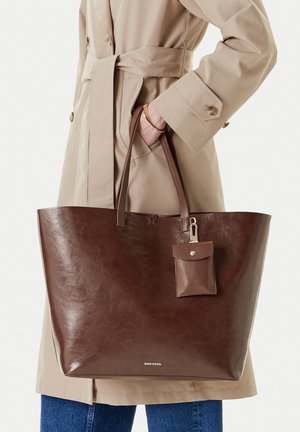 Persona con gabardina beige sosteniendo un gran bolso de cuero marrón con un pequeño bolso adjunto y detalles en oro.
