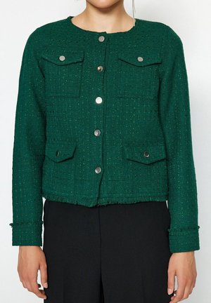 Femme portant une veste boutonnée texturée vert foncé avec quatre poches à rabat boutonnées, associée à un pantalon noir.