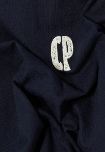 Tissu en coton bleu marine avec une texture froissée, arborant un logo "CP" brodé en fil blanc sur la partie supérieure gauche.