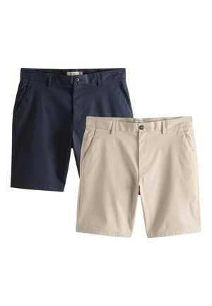 Mann trägt ein hellgraues Sweatshirt, ein weißes Unterhemd, schwarze Shorts, weiße Socken und weiße Turnschuhe und steht an einer schlichten Wand.
