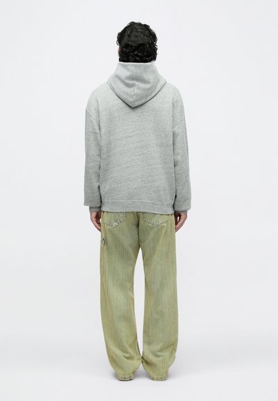 Uomo con capelli ricci scuri che indossa una felpa con cappuccio grigia e jeans verde sbiadito ampi, in piedi di fronte a uno sfondo bianco uniforme.