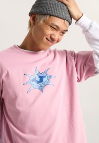 Camiseta de algodón rosa con un logo gráfico azul y un diseño de ráfaga en la parte frontal. Cuello redondo y mangas cortas.