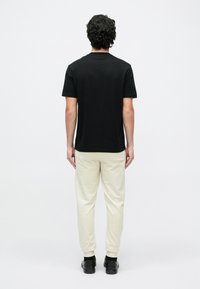 Göndör hajú férfi háttal áll, fekete pólót, krémszínű melegítőnadrágot és fekete cipőt visel, sima fehér háttér előtt.