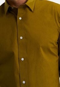 Oliivinvihreä, sileästä kankaasta valmistettu nappipaita. Siinä on klassinen kaulus ja kuusi valkoista nappia etukappaleen keskellä.