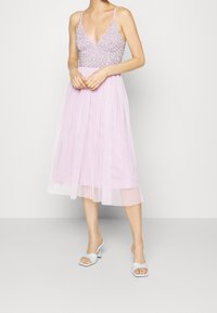 Mujer con un vestido midi sin mangas de color rosa claro, con un corsé de cuentas y una falda de gasa, combinado con sandalias de tacón blancas.