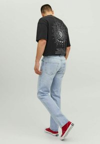 Camiseta gráfica negra, jeans de color azul claro y zapatillas rojas. La camiseta tiene un gran diseño en la parte posterior con texto y motivos circulares.