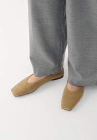 Tan semskede flate sko med firkantet tådesign og en lav svart såle, kombinert med vide grå bukser. Myk tekstur og minimalistisk stil.