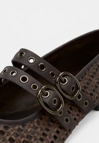 Zapato de cuero marrón trenzado con dos correas de cuero que presentan ojales de metal y hebillas ovaladas, visto en primer plano.