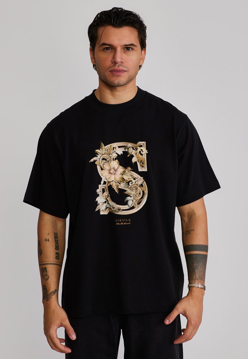 Homme aux cheveux foncés portant un T-shirt noir surdimensionné avec un grand motif floral doré orné en forme de "S" et plusieurs tatouages sur les avant-bras.
