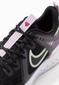 Chaussure de sport en mesh noire avec des accents lavande, présentant un détail en forme de cœur sur le laçage, et une semelle texturée blanche. Logo Nike visible.