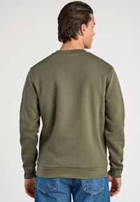 Sudadera verde oliva con cuello redondo, puños y dobladillo acanalados. Hecha de un tejido suave, mostrada desde la parte de atrás con jeans azules.
