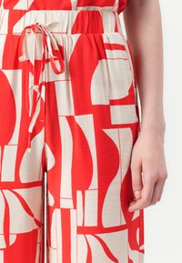 Vêtement à motif rouge et crème avec des formes abstraites, taille élastique avec cordon de serrage et poches latérales. Texture douce évidente.